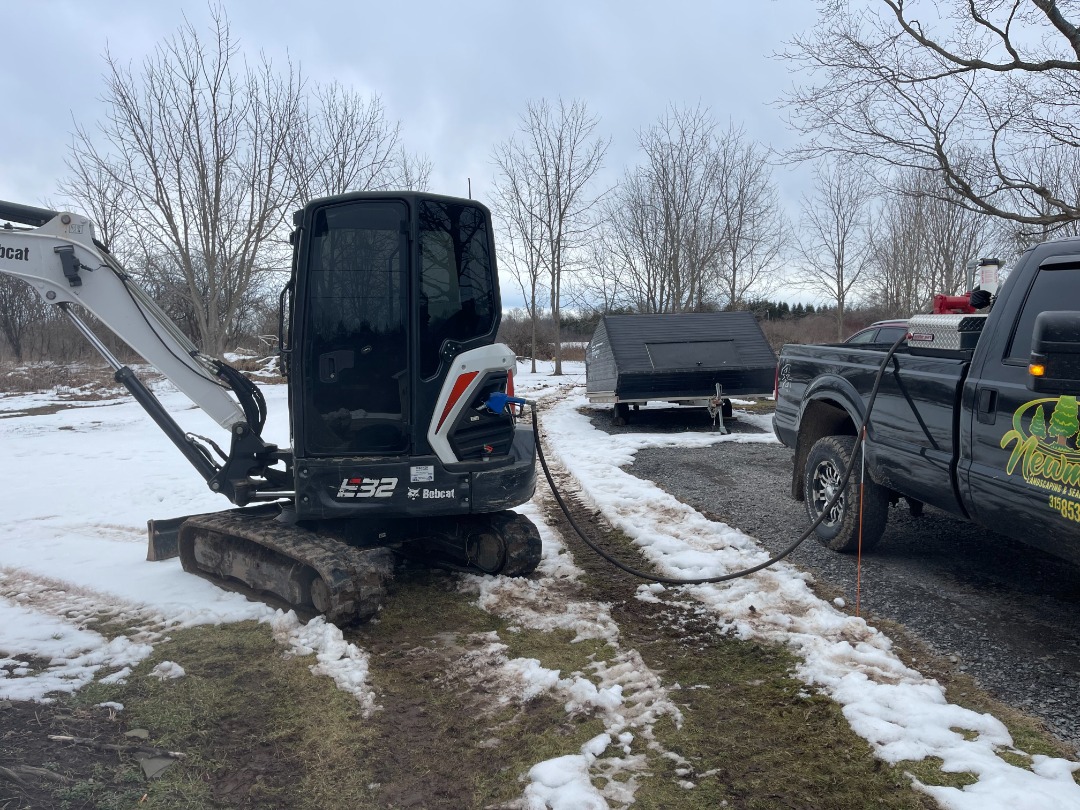 Bobcat E32 compact excavator being powered or towed by a landscaping truck on a snowy gravel lot during a cold winter day.
