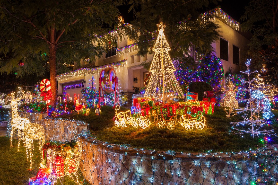 House covered in colorful Christmas lights, featuring glowing reindeer, candy canes, presents, and a large lit tree on the lawn.