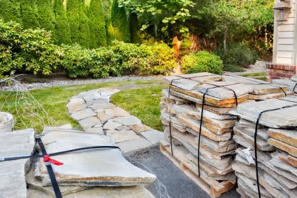 Flagstone walkway installation underway with stacked natural stone slabs near a curving garden path and green landscaping.