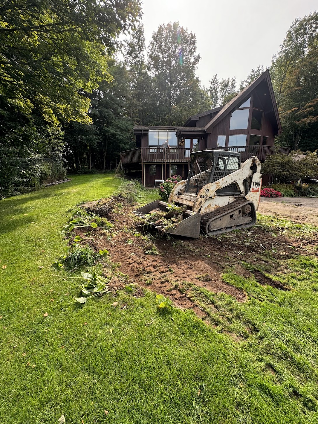 Bobcat T190 skid steer clears yard in front of modern cabin, starting excavation for landscape renovation near wooded area.