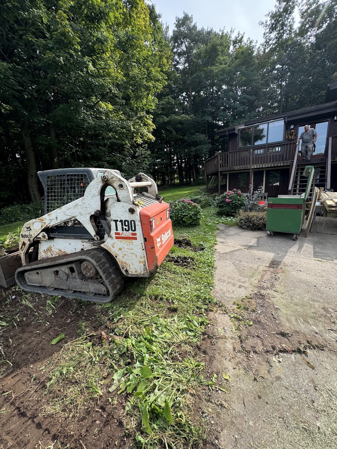 Skid steer prepares ground near house, removing brush and grass during residential excavation for new lawn installation.