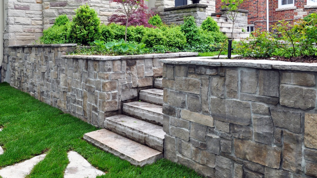 Stone retaining wall with built-in steps surrounded by lush green landscaping, creating terraced levels in a residential yard.