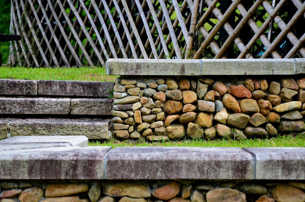 Rustic retaining wall made of rounded natural stones with stone slab steps, bordered by grass and a wooden lattice fence.