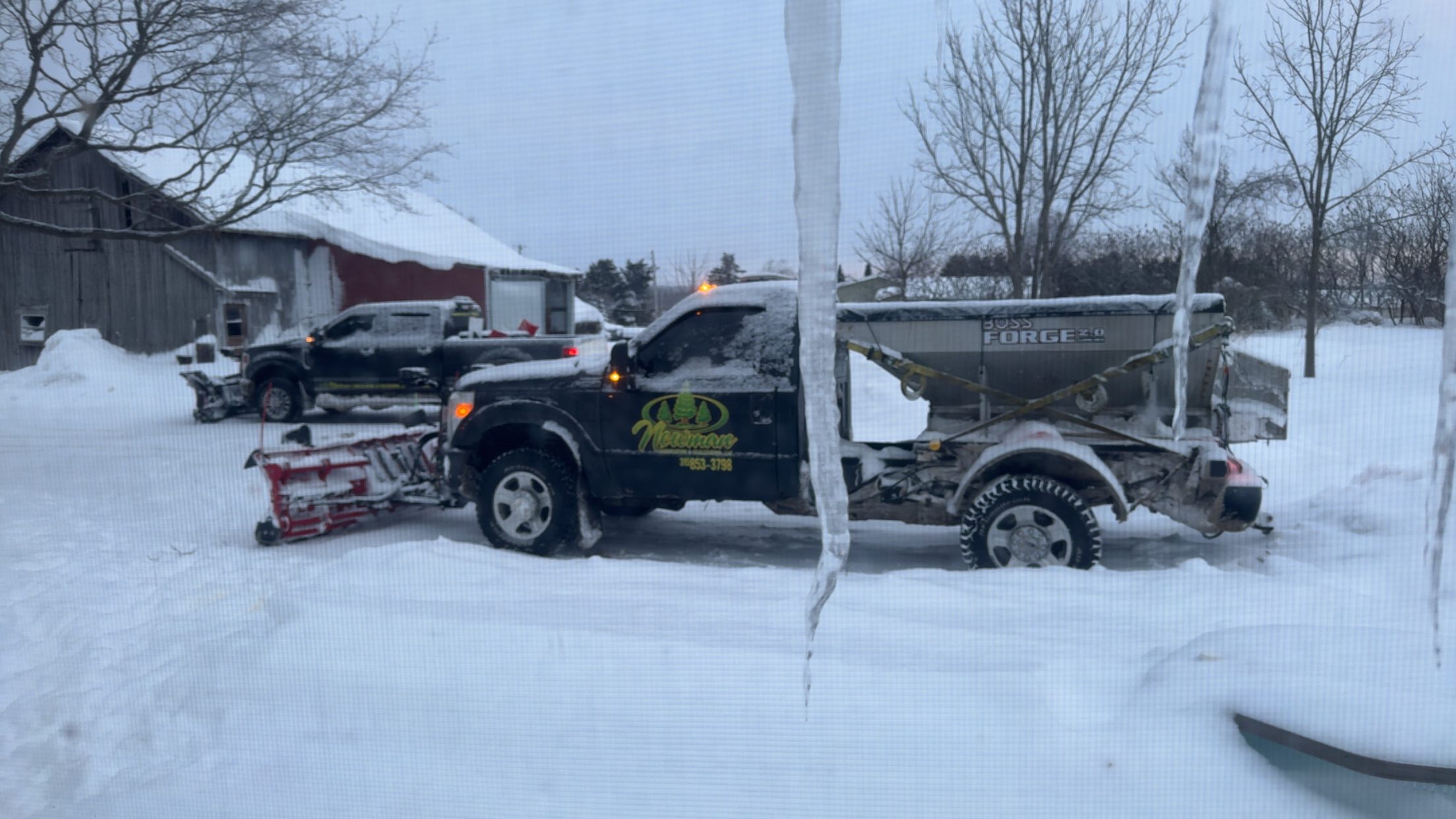 Snow removal truck with attached plow and salt spreader clearing residential driveways during a heavy winter snowstorm.