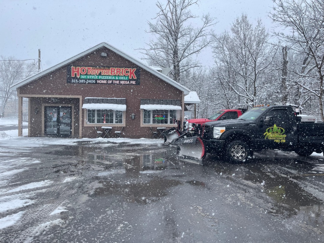  Snow removal truck parked in front of a pizzeria in Utica NY, ensuring safe business access during active snowfall.
