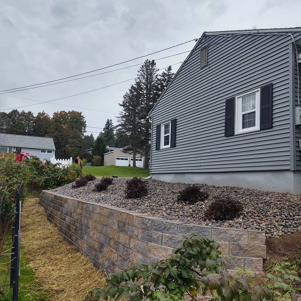 Stone retaining wall with rock drainage bed beside a gray house stabilizing a raised yard.